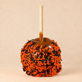 A Halloween-themed caramel apple with orange and black sprinkles, set against a solid beige background.