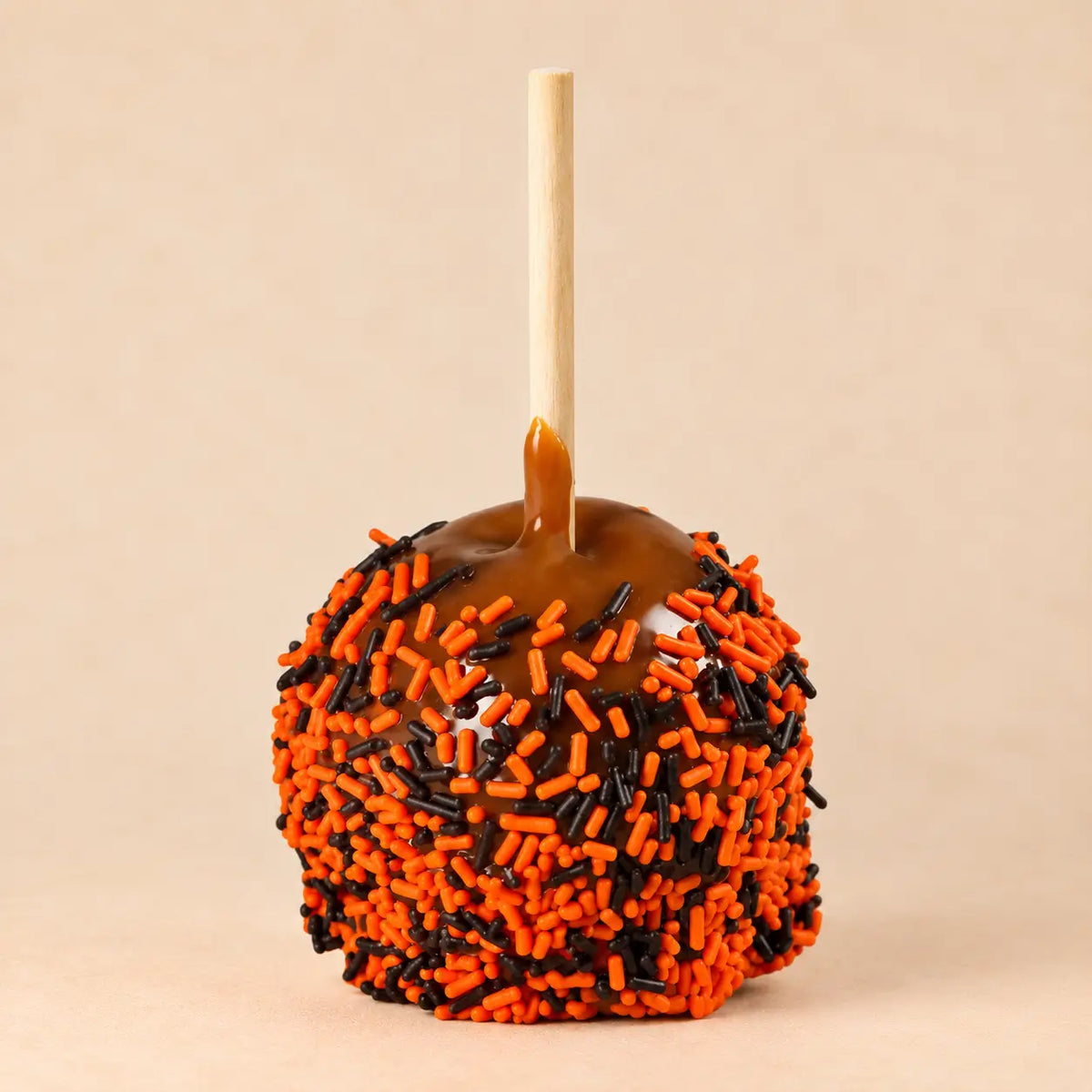 A Halloween-themed caramel apple with orange and black sprinkles, set against a solid beige background.