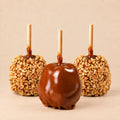 Three Morkes caramel apples placed on a warm beige background. The front apple is a plain caramel apple, and the two behind are peanut caramel apples.