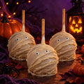 Three Halloween-themed caramel apple decorated like a mummy with white chocolate. They are set against a Halloween-themed background surrounded by Halloween decor.