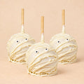 Three Halloween-themed caramel apple decorated like a mummy with white chocolate. They are arranged in a triangular form.