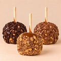 Three Snickers Caramel Apples arranged in a triangular form. In the front, there is the plain Snickers Caramel Apple. In the back, there are two Snickers Caramel Apples covered in milk and dark chocolate.